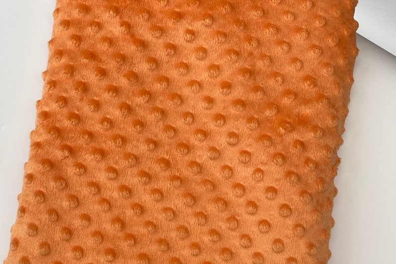 May include: Close-up of a soft, orange fabric with a textured, dotted pattern. The fabric is plush and has a fuzzy texture.