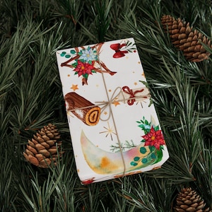 May include: A wrapped gift with a festive design featuring a crescent moon, stars, and floral elements in red, blue, and green. The gift is tied with twine and rests on a bed of green pine needles, with pine cones in the background.
