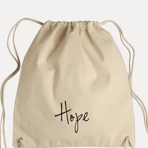 Op de afbeelding: Een beige trekkoordtas met het woord "Hope" in zwarte cursieve letters op de voorkant.