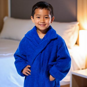 May include: A young person wearing a vibrant blue terry cloth robe with a shawl collar and pockets. The robe is a solid, bright blue color. The person is smiling, and the background includes a bed and a lamp.