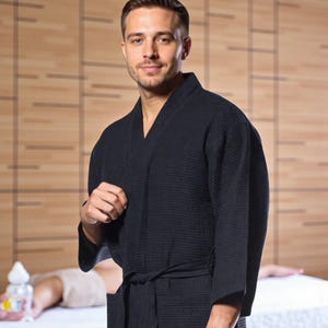 May include: A black waffle-weave robe with a matching tie. The robe has a shawl collar and long sleeves. The person in the image is wearing the robe. The background is a spa setting with wooden paneling.
