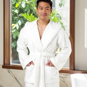 May include: A white hooded bathrobe with a matching belt and pockets. The plush robe is made of a soft, absorbent material. The person is standing in front of a window with green foliage visible in the background.