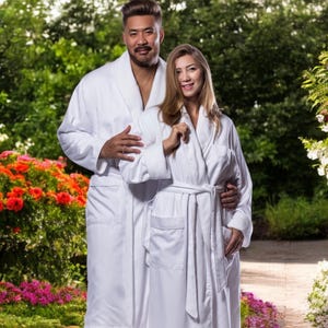 May include: Two people wearing white bathrobes, standing in a garden setting with green trees and flowers.