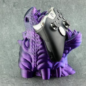 May include: A black video game controller is held by a purple monster-themed stand. The stand is 3D-printed, with a textured, scaly design and sharp teeth. The controller has silver accents. The stand is purple.