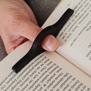 Könnte beinhalten: Ein schwarzer, ergonomischer Daumen-Seitenhalter aus strukturiertem Material. Das geschwungene Design ermöglicht bequemes einhändiges Lesen. Dieses Lesezubehör hilft, Seiten offen zu halten.