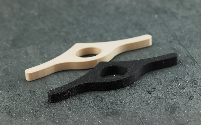 May include: Two book holders, one beige and one black, are displayed on a textured gray surface. Each holder has a curved design with a circular opening. The black holder has a speckled appearance.