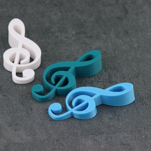 May include: Three treble clef music note shapes, one white, one teal, and one light blue, on a grey textured surface.