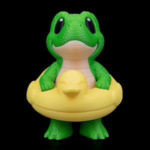 以下が含まれることがあります： 大きな黒い目と笑顔の表情をした緑色のカエルの置物。カエルはアヒルのデザインの黄色い浮き輪を身に着けています。置物は黒い背景に配置されています。