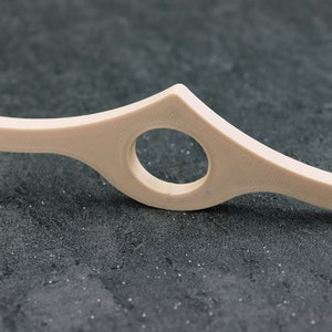 May include: A beige, curved book page holder with a circular opening in the center. The holder is designed to keep a book open and is made of a smooth, solid material. The design is simple and functional.