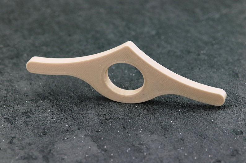 May include: A beige, curved book page holder with a circular opening in the center. The holder is designed to keep a book open and is made of a smooth, solid material. The design is simple and functional.