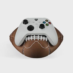 Op de afbeelding: Een witte Xbox-controller ligt bovenop een bruine American football. De controller heeft zwarte joysticks en gekleurde knoppen. De voetbal is bruin met witte stiksels.