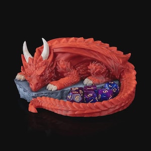 Può includere: Una statuina di drago rosso con corna e artigli bianchi, appoggiata su una base di pietra grigia. Diversi dadi poliedrici viola e blu sono raggruppati vicino al drago, suggerendo un tema di gioco da tavolo.