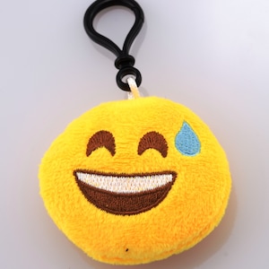 Peut inclure: Porte-clés en peluche jaune avec un visage d'emoji souriant et une larme bleue. Le porte-clés a un clip en plastique noir.