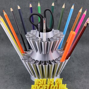 May include: A silver metal pencil holder with multiple slots for pencils, pens, and other art supplies. The holder has two tiers and is filled with colorful pencils, pens, and a pair of scissors. The holder has a yellow base with the text "VISIT US ON 3DGIFT.CO.UK".