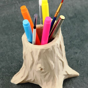 Könnte beinhalten: Ein beigefarbener, baumstumpf-förmiger Stiftehalter mit einer Vielzahl von bunten Stiften und Markern im Inneren. Der Halter hat eine raue, strukturierte Oberfläche.