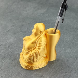Könnte beinhalten: Goldfarbene Figur eines lächelnden Buddha, der einen Geldbeutel hält, als Stifthalter verwendet. Die Figur steht auf einer grauen Oberfläche.