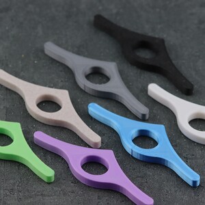 May include: A collection of colorful, 3D-printed book page holders. The holders are in various colors, including green, purple, blue, gray, black, white, and beige. Each has a circular opening for the finger and a curved design to hold pages open.