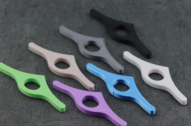 May include: A collection of colorful, 3D-printed book page holders. The holders are in various colors, including green, purple, blue, gray, black, white, and beige. Each has a circular opening for the finger and a curved design to hold pages open.