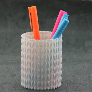 May include: A white ceramic pencil holder with a textured pattern, holding three markers: one orange, one pink, and one blue.