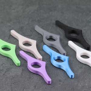 May include: A collection of seven colorful thumb page holders in various shades, including green, beige, purple, blue, gray, black, and white. These ergonomic reading accessories are designed to hold book pages open.