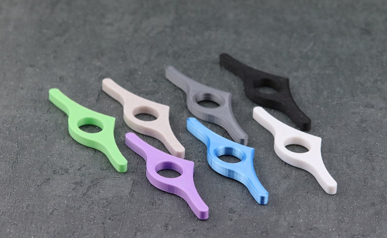 May include: A collection of seven colorful thumb page holders in various shades, including green, beige, purple, blue, gray, black, and white. These ergonomic reading accessories are designed to hold book pages open.
