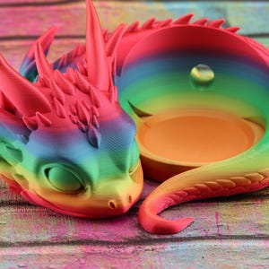 Peut inclure: Un pot de fleurs en forme de dragon aux couleurs de l'arc-en-ciel avec un petit trou en haut. Le pot est fait d'un matériau lisse, semblable au plastique, et a une queue enroulée.