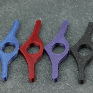 May include: Four colorful book page holders in blue, red, purple, and black. Each has a curved handle and a circular opening. The objects are made of a textured material and are arranged on a gray surface.