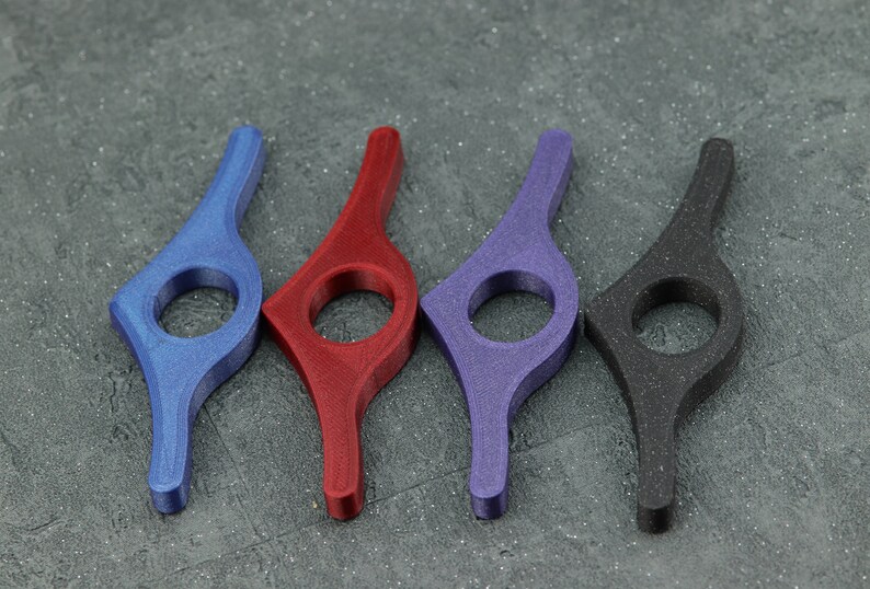 May include: Four colorful book page holders in blue, red, purple, and black. Each has a curved handle and a circular opening. The objects are made of a textured material and are arranged on a gray surface.