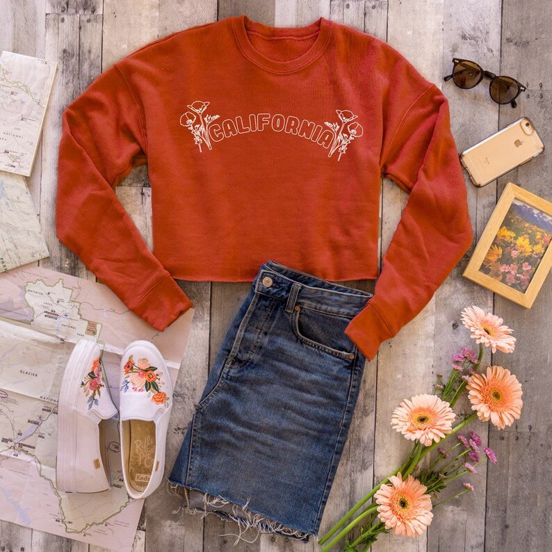 May include: A rust-colored cropped sweatshirt with the text "CALIFORNIA" in white with floral accents. The sweatshirt is paired with a denim skirt and white sneakers with floral embroidery.
