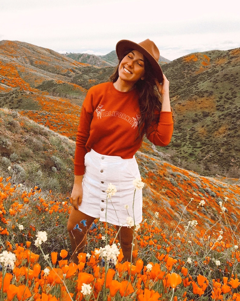 May include: A woman wearing a red sweatshirt with the word "CALIFORNIA" embroidered on it, a white skirt, and a brown hat, standing in a field of orange poppies.