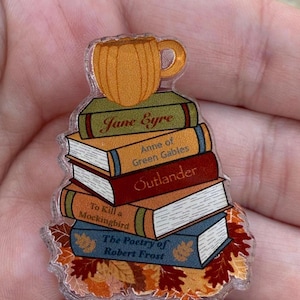 Pode incluir: Um pin de acrílico transparente com uma pilha de livros, incluindo "Jane Eyre" e "Outlander", encimada por uma caneca laranja. O pin tem um tema de outono com folhas de outono na base.