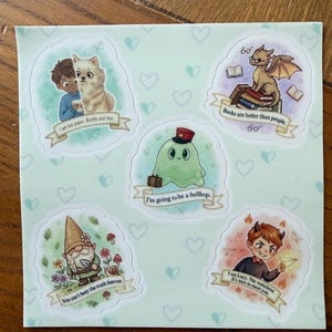 Puede incluir: Una hoja de pegatinas con ilustraciones caprichosas. Los diseños incluyen un niño y un gato, un dragón sobre libros, un fantasma verde, un gnomo y un niño con cuernos. Cada pegatina tiene una cita única.