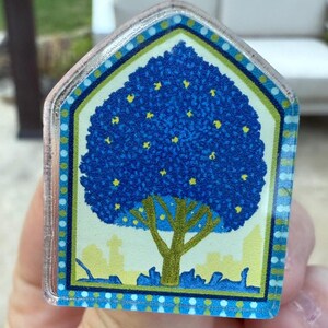 May include: A clear, house-shaped acrylic magnet featuring a stylized blue tree with yellow accents. The tree is set against a light background with a cityscape at the base, framed by a blue and green border.