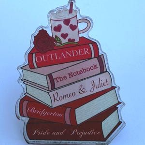 May include: A clear acrylic pin shaped like a stack of books with titles including "Outlander", "The Notebook", "Romeo & Juliet", "Bridgerton", and "Pride and Prejudice". A coffee cup with heart designs and a red rose are on top.