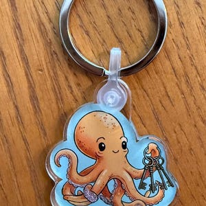 May include: A keychain featuring a cartoon octopus holding keys. The octopus is orange with a light blue background. The keychain has a silver ring.