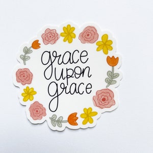 Puede incluir: Una pegatina blanca con un diseño de corona de flores. La corona está hecha de flores rosas y amarillas y tulipanes naranjas. El texto "grace upon grace" está escrito en cursiva negra dentro de la corona.