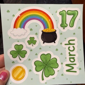 May include: Sticker sheet with St. Patrick's Day themed graphics. Includes a rainbow, pot of gold, shamrocks, a gold coin, and the number 17. The word "March" is also present. The stickers have a white border and are on a light green background.