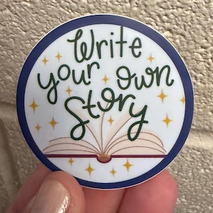 May include: A round sticker with a white background and a navy blue border. The sticker features the text "Write your own story" in green script, with an open book illustration and gold star accents.