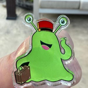 May include: A clear, glossy pin featuring a cartoon green alien wearing a red and black bellhop hat. The alien is smiling and holding a brown suitcase. The pin has a clear border.
