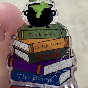 May include: A clear acrylic pin featuring a stack of books with titles like "Frankenstein" and "Gothic Horror." A black cauldron with green bubbling liquid sits atop the books. A white spiderweb design is on the side of the books.