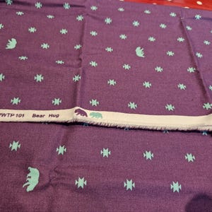 Pode incluir: Tecido roxo com um padrão repetido de ursos turquesa e formas geométricas. Uma faixa branca com o texto "PINK PWTP 101 Bear Hug" é visível. Adequado para projetos de costura ou artesanato.