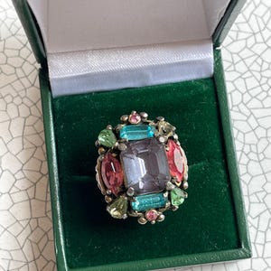 May include: A vintage ring featuring a large, rectangular, grey-purple central stone, encircled by smaller gems in pink, green, and blue. The ring is set in gold-toned metal and displayed in a green velvet-lined box.