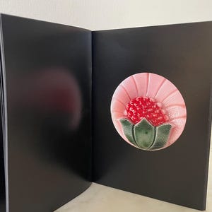 May include: A black open book or folder featuring a circular ceramic inlay on the right side. The inlay displays a stylised pink flower with a textured red centre and green leaves at the base, set against a pale pink radiating background.