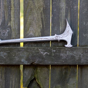 May include: A silver-colored, decorative axe with an ornate design. The axe features a long, slender handle and a detailed head with curved edges and decorative patterns. The axe is resting on a weathered wooden fence.