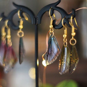 May include: Earrings with a delicate, translucent wing design, displayed on a black stand. The wings feature gold accents and are suspended from gold-colored hooks. The earrings have a whimsical, fairy-like aesthetic.