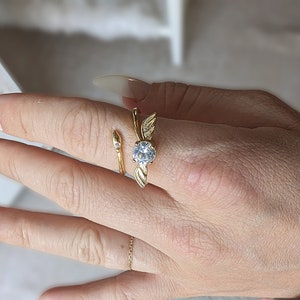 Peut inclure: Une bague dorée avec une grosse pierre précieuse transparente au centre. La bague présente un motif en forme de feuille de chaque côté de la pierre précieuse et une petite pierre précieuse sur la bande. La bague est sur le doigt d'une personne.