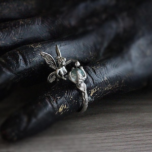 May include: A silver ring featuring a fairy holding a gemstone. The fairy has detailed wings and is positioned next to a round, light blue gemstone. The ring is set against a dark background.