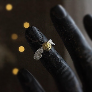 May include: A gold and silver ring in the shape of a winged ball, set against a dark background. The ring features a detailed design with silver wings and a gold-colored body. The ring is on a black finger.