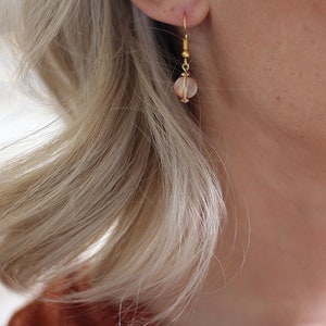 May include: A close-up of a gold earring with a small, round, translucent bead. The earring is attached to a person's ear. The person has blonde hair and fair skin. The earring is the focal point of the image.