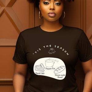 May include: A brown t-shirt with a white graphic design that says "'Tis the season" with a bell, a pepper pot, a slice of black cake, and a loaf of bread. The text below the graphic says "Pepper Pot", "Black Cake", and "Home-made Bread".